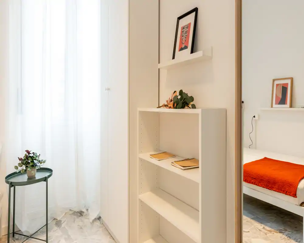 Rincón de dormitorio moderno y luminoso visible a través de una puerta abierta, cama blanca con una manta roja cálida y decoración minimalista; la luz natural a través de la cortina crea un ambiente ligero.