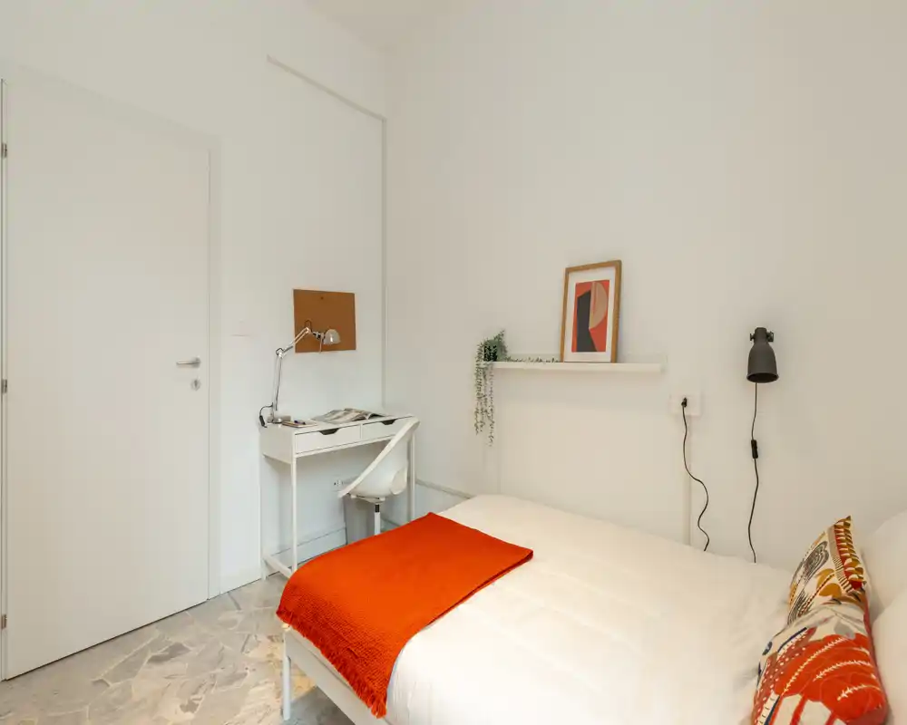 Dormitorio luminoso y minimalista con una cama doble cómoda con ropa blanca y una manta naranja; un pequeño escritorio y silla junto a la pared, decoración sencilla y moderna.