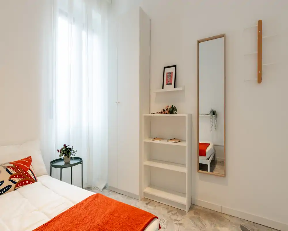 Dormitorio luminoso y minimalista con paredes blancas, cama acogedora con manta roja y cojines estampados, armario, estantes y espejo de pie — perfecto para un anuncio de alquiler moderno.