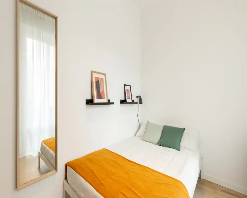 Dormitorio minimalista y luminoso con una cama doble cómoda, cojines verdes y una cálida manta naranja. Un espejo alto y cuadros sencillos en la pared aportan profundidad y un aspecto moderno.