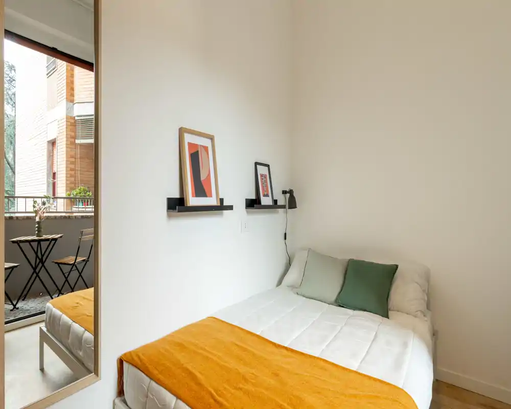 Dormitorio compacto y luminoso con cama bien hecha, cojines verdes y una cálida manta mostaza. Un espejo de cuerpo entero y un balcón con conjunto de bistro aportan profundidad y luz natural.