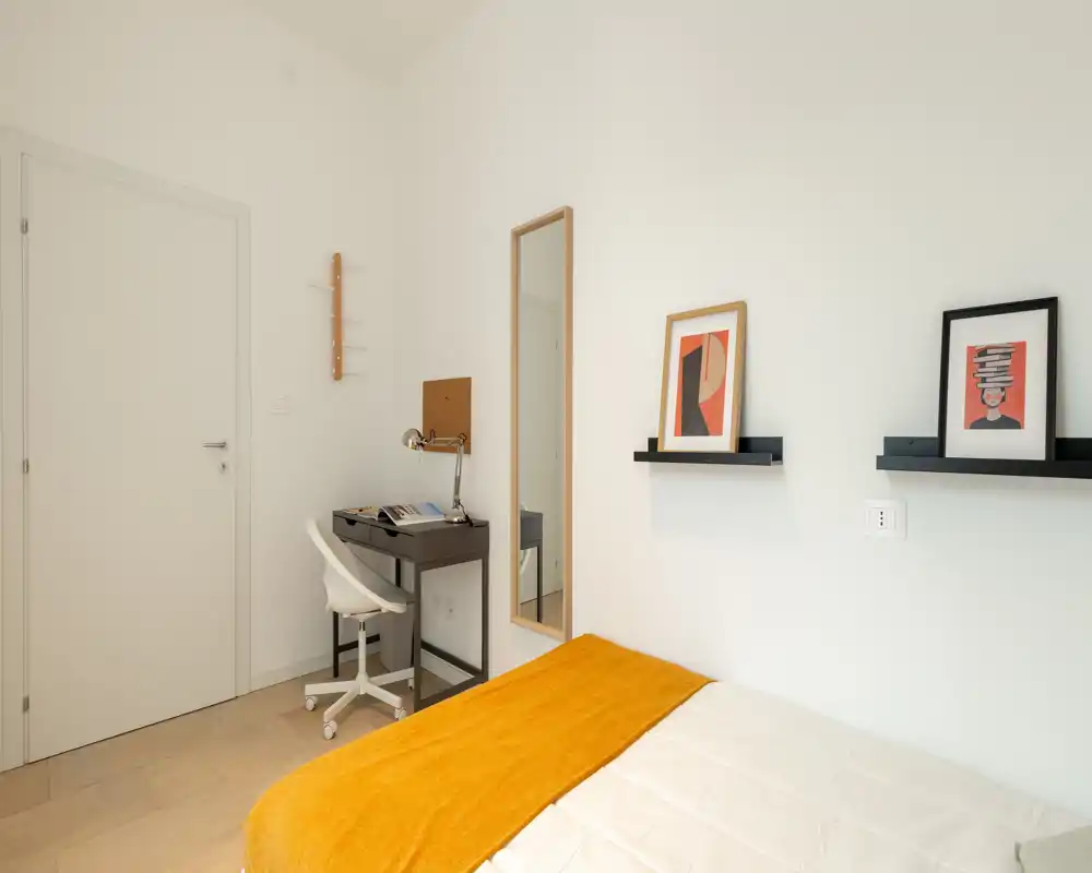Dormitorio luminoso y minimalista con cama cómoda y una manta naranja cálida, pequeño escritorio y espejo—ideal para una persona o estancias cortas.