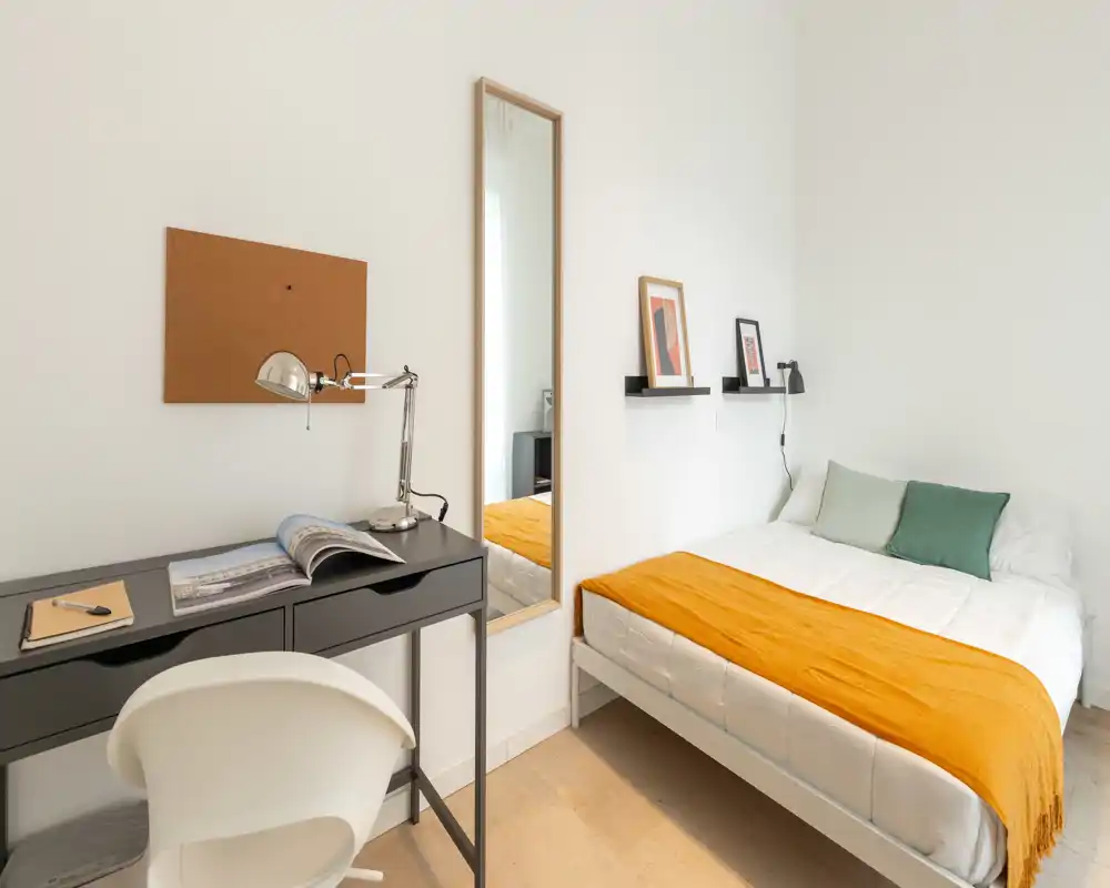 Dormitorio luminoso y minimalista con cama bien hecha y una manta naranja, un pequeño escritorio con lámpara y un espejo de cuerpo entero — ideal para un alquiler compacto y con estilo.