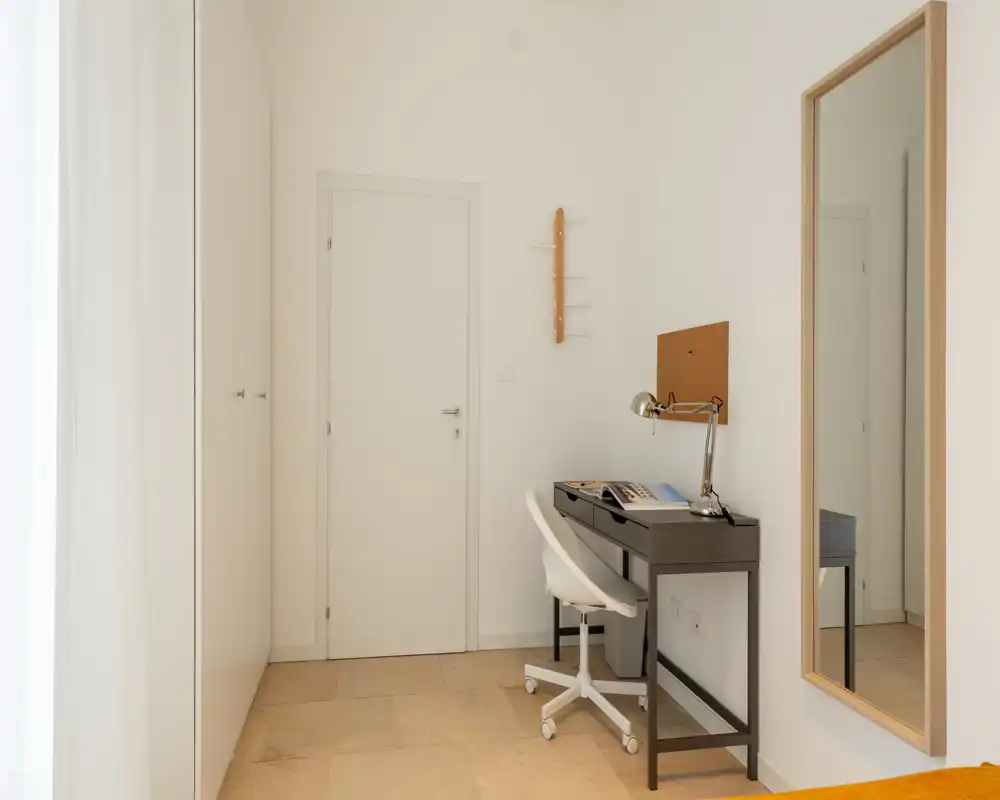 Entrada luminosa y minimalista con un pequeño escritorio y un espejo. Tonos neutros, suelo de baldosas y armario empotrado que crean un acceso limpio y funcional.