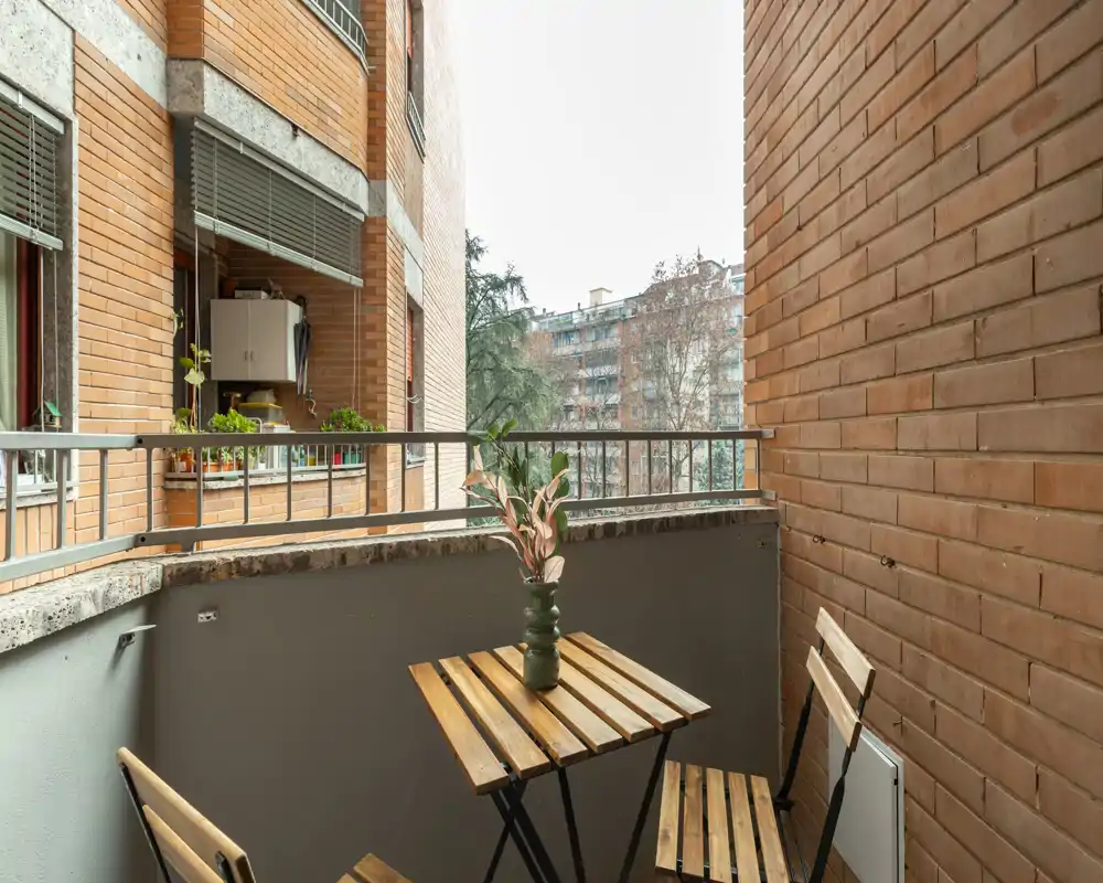 Pequeño balcón urbano con mesa bistró de madera y dos sillas, paredes de ladrillo y una planta en maceta; ideal para tomar un café por la mañana con vista a la ciudad.