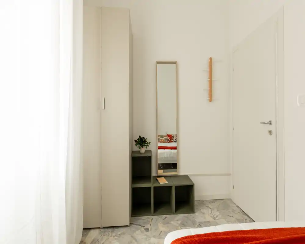 Rincón de dormitorio luminoso y minimalista con armario, espejo de pie y banco modular; una manta roja en la cama aporta un detalle cálido visible en la parte inferior.