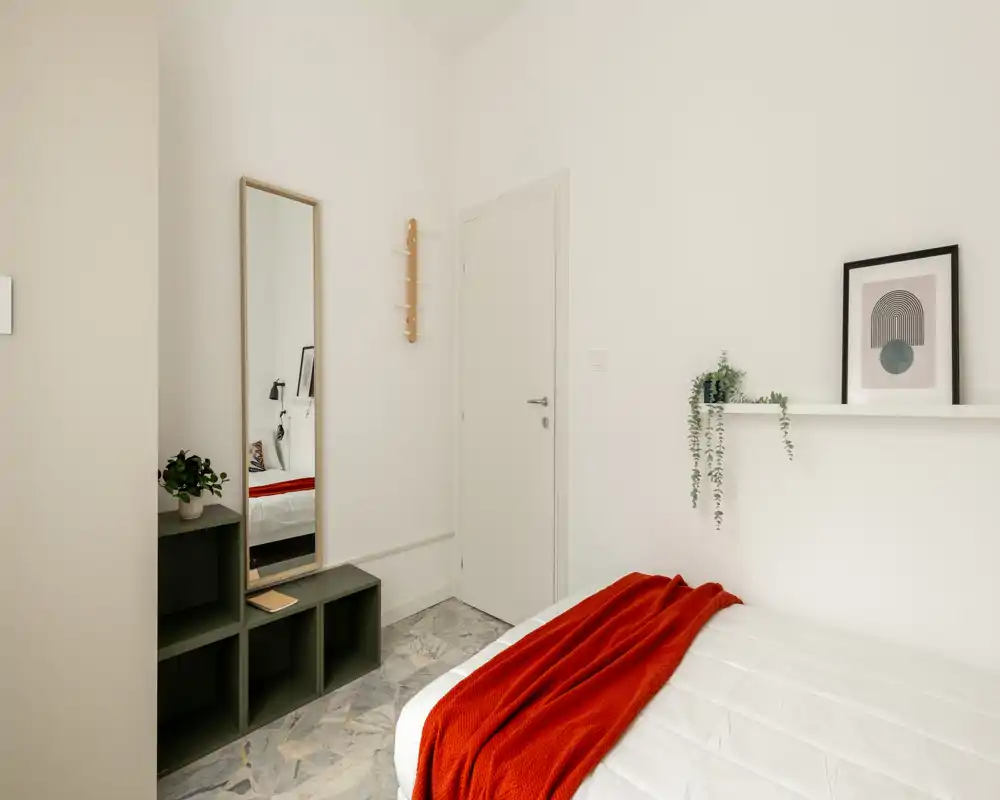 Dormitorio luminoso y minimalista con cama bien hecha, manta roja, estantería con cuadro y planta colgante, y espejo de cuerpo entero junto a la puerta.