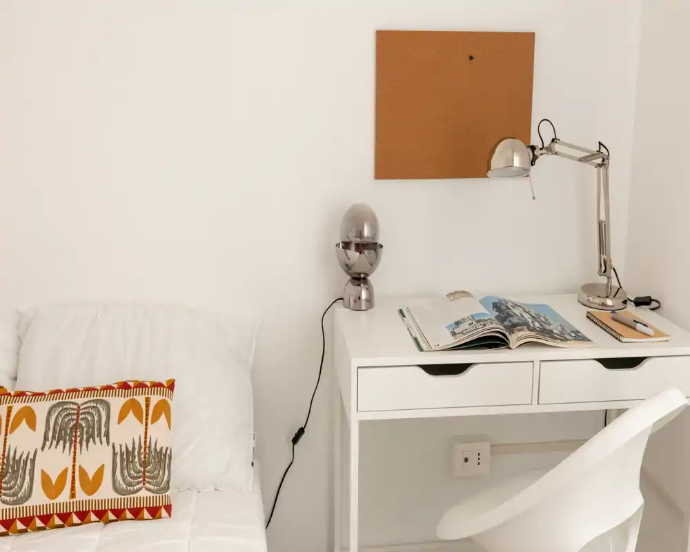 Rincón de dormitorio luminoso y minimalista con cama blanca y pequeño escritorio blanco con lámpara y libro abierto — ideal para estudios o habitaciones de invitados.