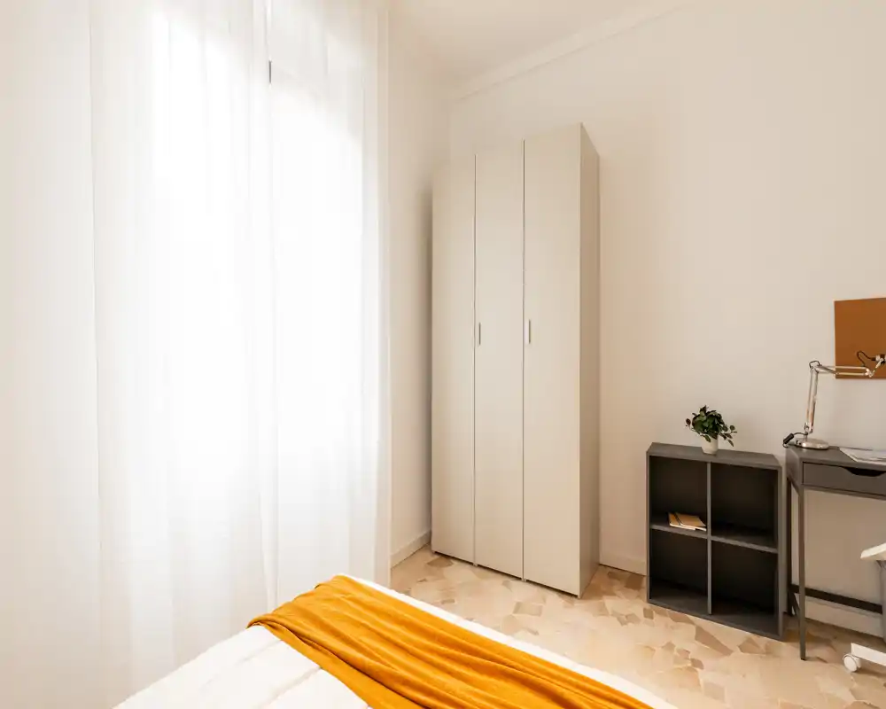 Rincón de dormitorio luminoso y minimalista con armario blanco, estantería pequeña y cama bien hecha con una manta naranja; luz natural suave a través de cortinas translúcidas.