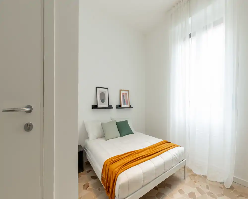 Dormitorio luminoso y minimalista con cama doble bien hecha, ropa blanca, cojines verdes y una manta naranja junto a una gran ventana con abundante luz natural.