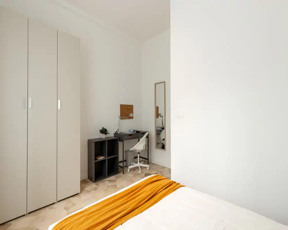 Dormitorio luminoso y minimalista con cama visible vestida de blanco y una manta naranja, armario y un escritorio compacto — ordenado y funcional para viajeros o estudiantes.