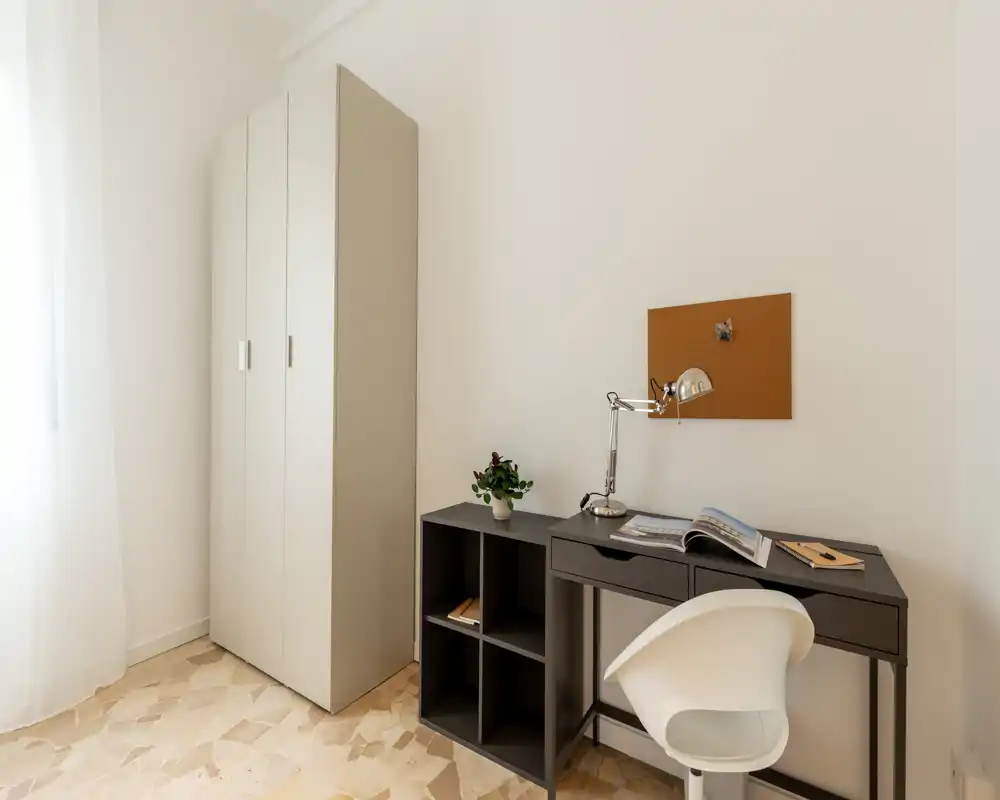 Rincón de dormitorio luminoso y minimalista con un armario alto, un pequeño escritorio y silla, y un corcho encima del área de trabajo — ideal para un estudio compacto.