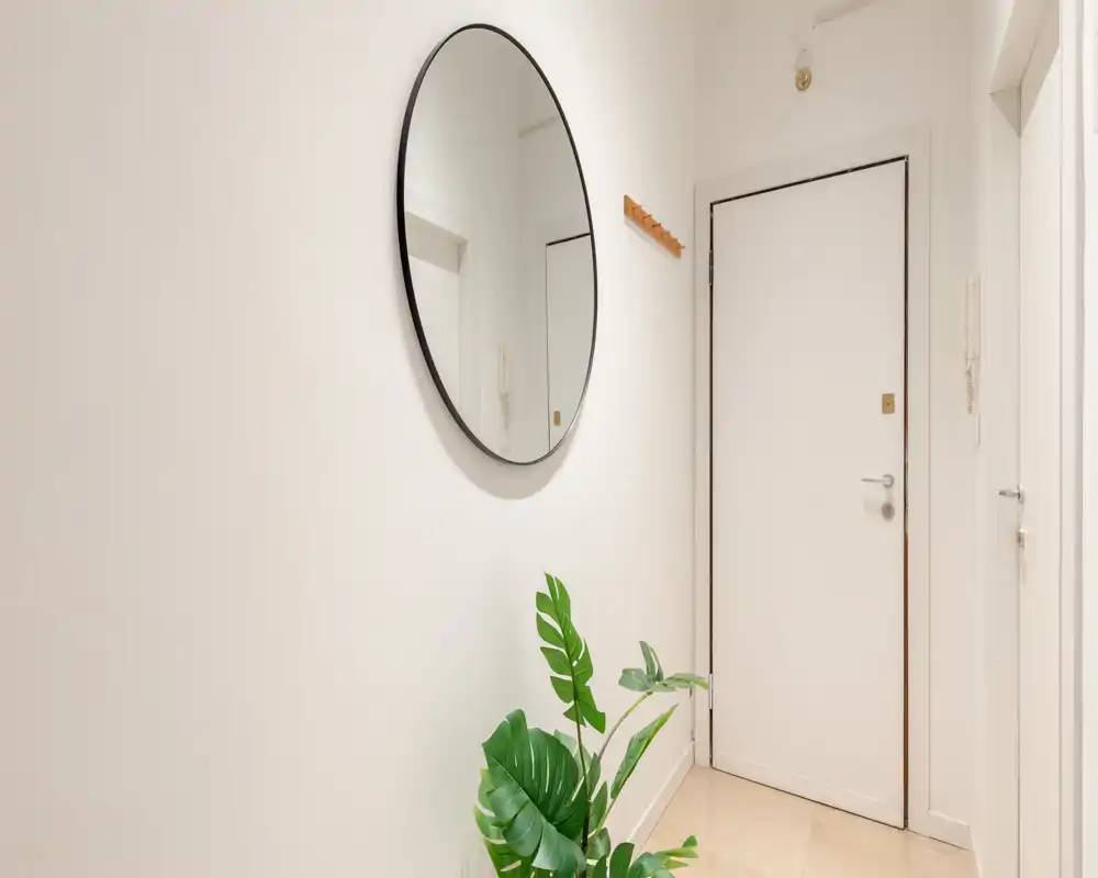 Entrada luminosa y minimalista con un espejo redondo en la pared, puerta blanca y planta decorativa, que ofrece una primera impresión limpia y acogedora.