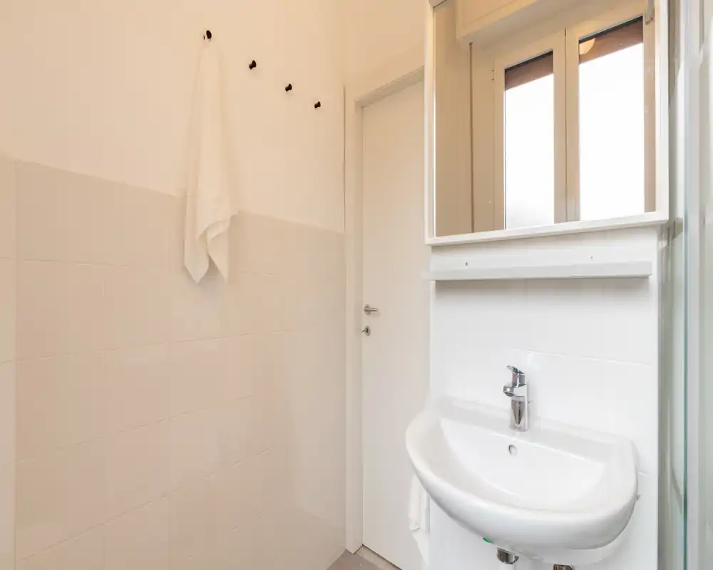 Baño compacto y limpio con azulejos blancos, lavabo mural, espejo con armario y gancho para toalla; diseño luminoso y minimalista ideal para un piso pequeño.
