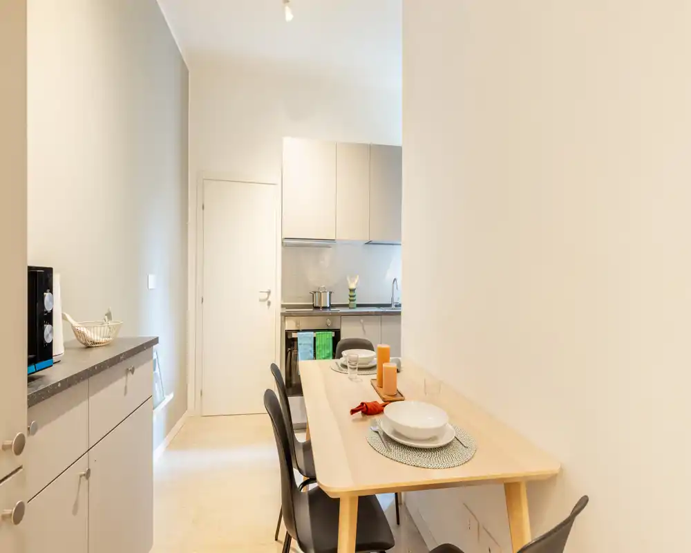 Área de comedor compacta junto a una cocina moderna, con mesa de madera clara puesta con manteles, velas y cuatro sillas estilizadas: luminosa, minimalista y lista para comer.