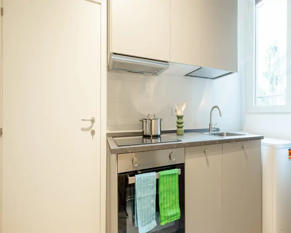 Cocina compacta moderna con horno integrado, placa de inducción y fregadero junto a una ventana luminosa — bien presentada con paños de colores y una olla en la placa.