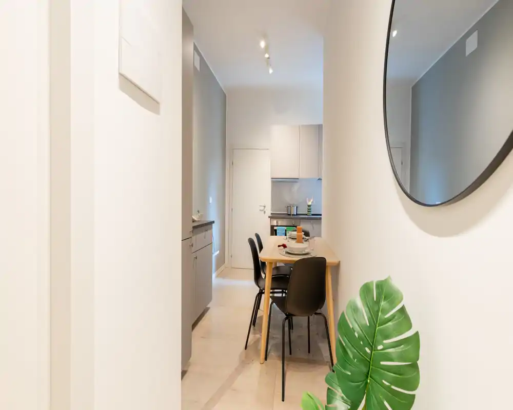 Área de comedor moderna y luminosa con una mesa de madera pequeña puesta para comer, cuatro sillas negras y vista a la cocina compacta — líneas limpias y decoración cuidada que crean un ambiente acogedor.