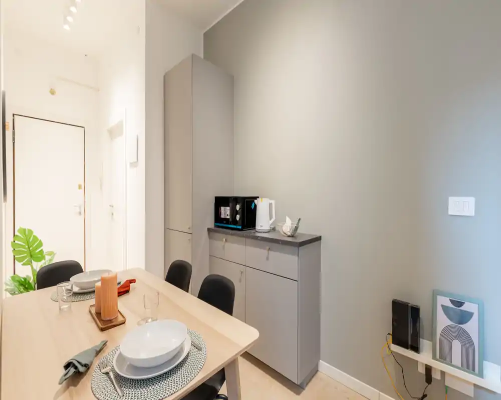 Zona de comedor bien decorada con mesa de madera clara puesta para dos, sillas cómodas y un armario auxiliar con pequeños electrodomésticos. Tonos neutros e iluminación suave crean un rincón comedor moderno y acogedor.