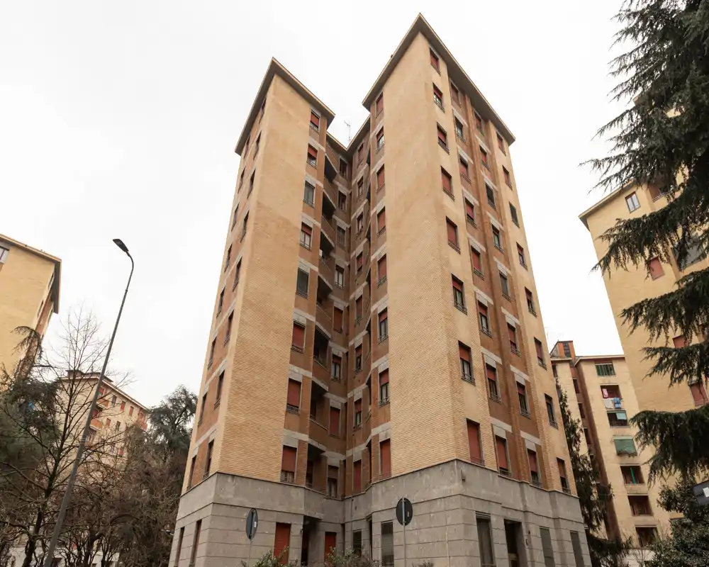 Vista exterior de un edificio de apartamentos de varios pisos en ladrillo con múltiples ventanas y base de piedra, enmarcado por árboles y señales de tráfico.