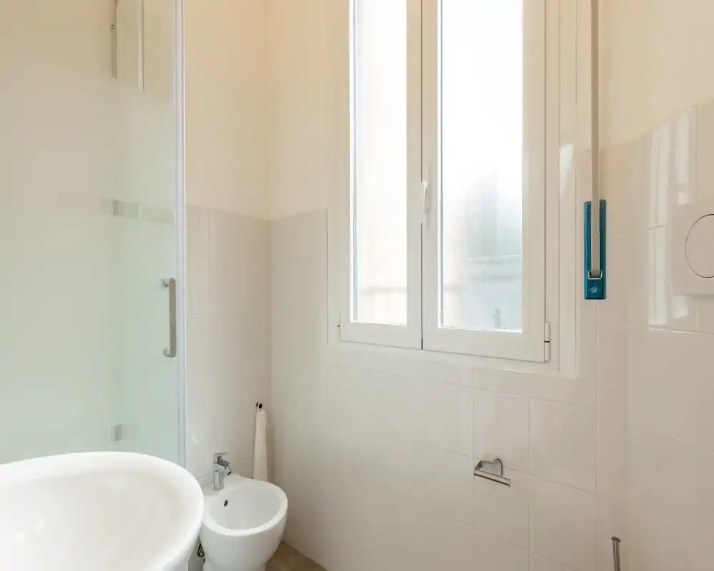 Baño pequeño y luminoso con lavabo blanco, bidé y ducha de vidrio esmerilado; paredes alicatadas y ventana que aporta luz natural.