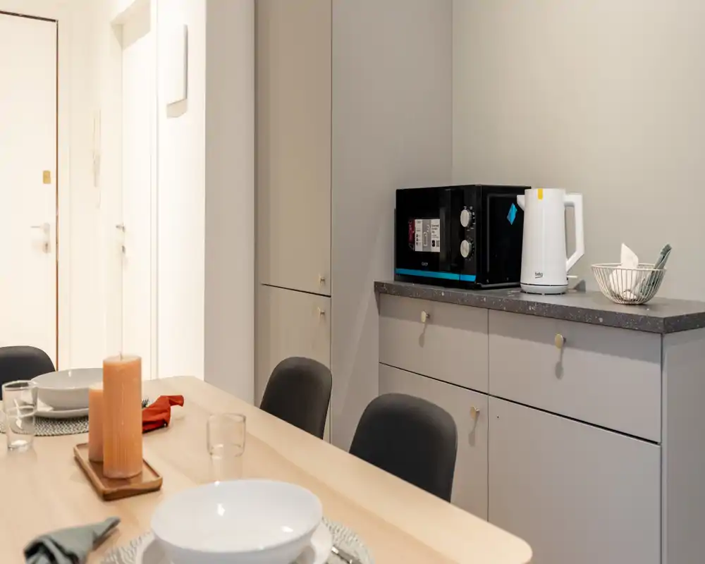 Área de comedor bien decorada con mesa de madera montada para comer, sillas cómodas y armarios empotrados; un microondas y una tetera sobre la encimera.
