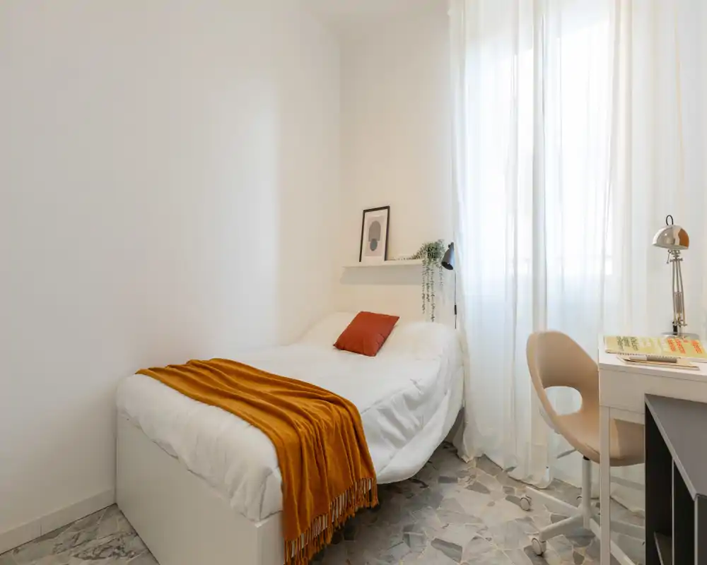 Camera da letto luminosa e minimal con un singolo letto ordinato, una coperta calda e una piccola scrivania accanto a una grande finestra che diffonde luce naturale.