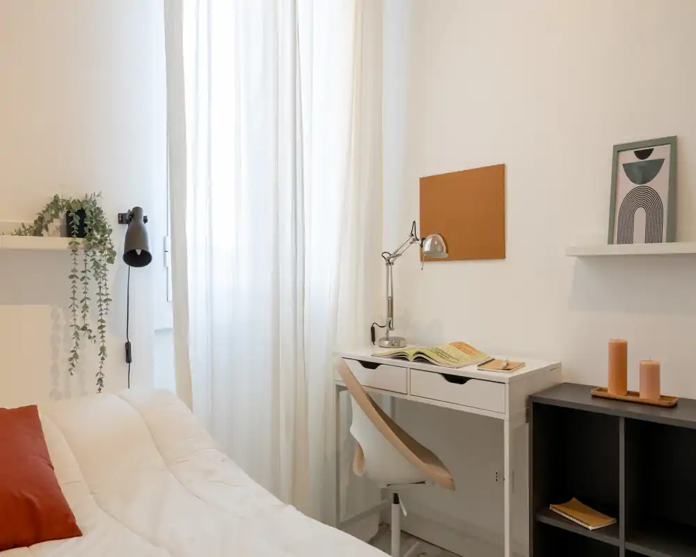 Camera da letto luminosa e minimal con letto bianco, tessuti morbidi e una piccola scrivania vicino alla finestra — perfetta per una stanza singola accogliente.