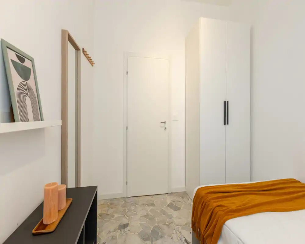 Camera da letto luminosa e minimal con letto ordinato coperto da un caldo plaid arancione, un armadio bianco alto e dettagli decorativi discreti che creano un’atmosfera moderna e rilassante.
