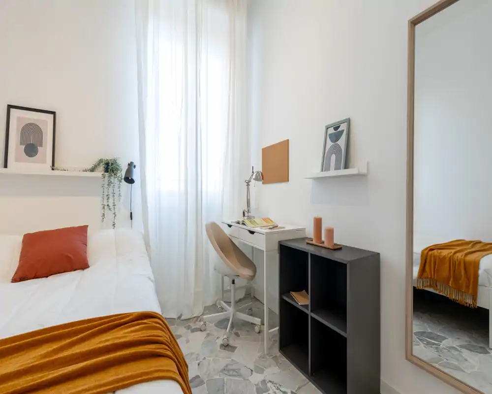 Camera da letto luminosa e arredata con un comodo letto bianco, plaid e cuscino arancioni, una scrivania sotto la finestra e uno specchio a figura intera che aumenta la profondità.