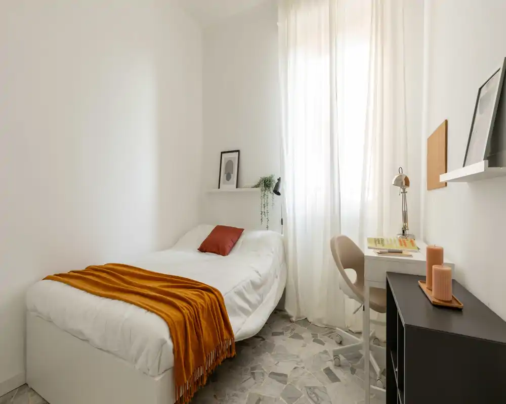 Camera singola luminosa e minimal con letto bianco, plaid arancione caldo e una scrivania vicino alla finestra soleggiata.