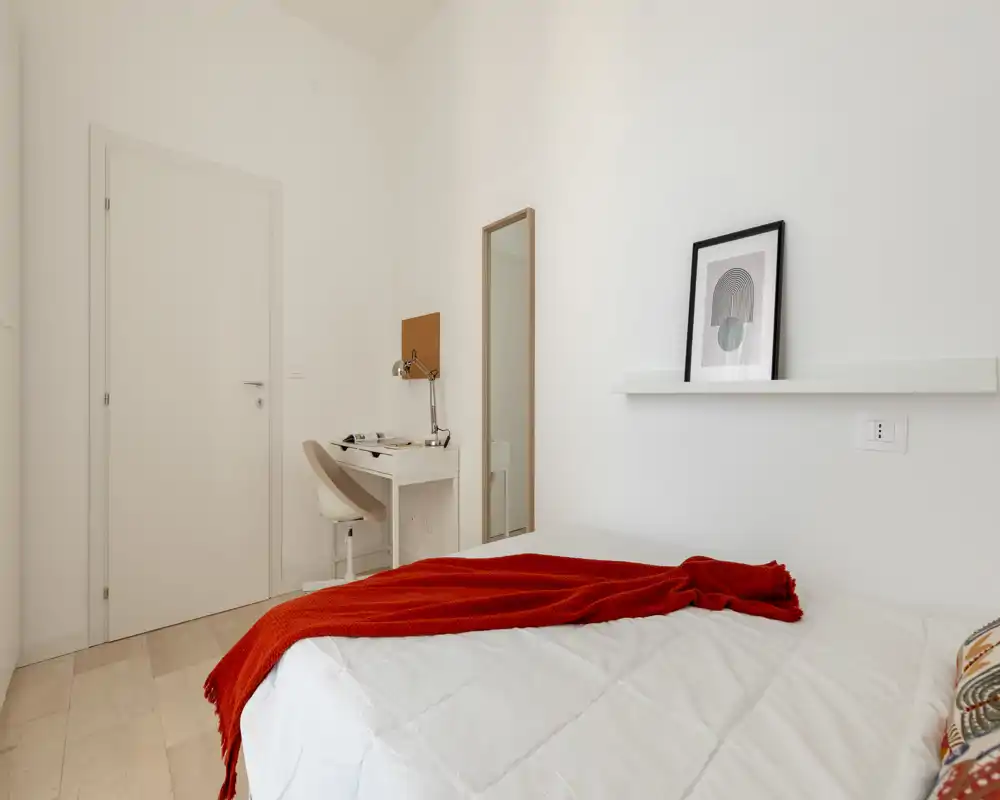 Camera da letto minimalista e luminosa con piumone bianco e plaid rosso, piccolo scrittoio con lampada e specchio a figura intera — ambiente pulito e accogliente, ideale per una persona.