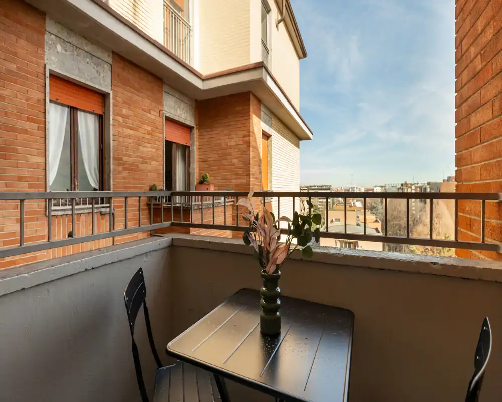 Piccolo balcone soleggiato con tavolino in metallo e due sedie, affacciato su edifici in mattoni e vista cittadina in lontananza.