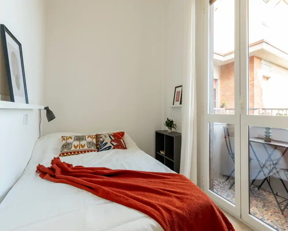 Piccola camera da letto luminosa con letto bianco, cuscini decorativi e una coperta color ruggine; ampia finestra/porta sul balcone che garantisce molta luce naturale.