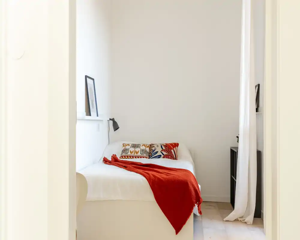 Camera da letto luminosa e minimal con letto bianco, cuscini decorativi colorati e un plaid rosso che crea un accento caldo. Mensola semplice, lampada da comodino e tende leggere completano l'aspetto moderno e tranquillo.