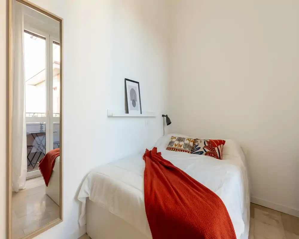 Camera da letto luminosa e minimale con letto matrimoniale con lenzuola bianche, cuscini decorativi e un caldo plaid rosso, illuminata dalla porta sul balcone.