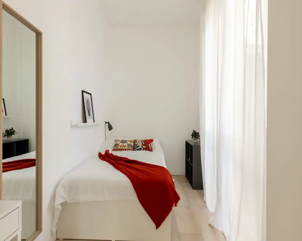 Camera da letto luminosa e minimal con un letto singolo ben rifatto, cuscino decorativo e plaid rosso, luce naturale da una grande finestra — perfetta per un monolocale accogliente.