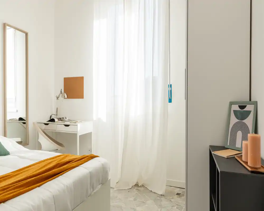 Bright, minimalist bedroom with a neatly made bed, sheer curtains letting in soft natural light, a small desk and decorative accents for a calm, modern feel.