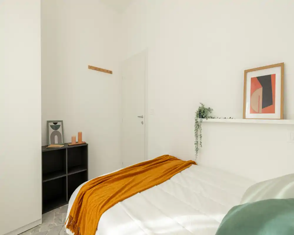 Bright, minimal bedroom with a neatly made bed, warm ochre throw and simple decor creating a calm, modern feel.