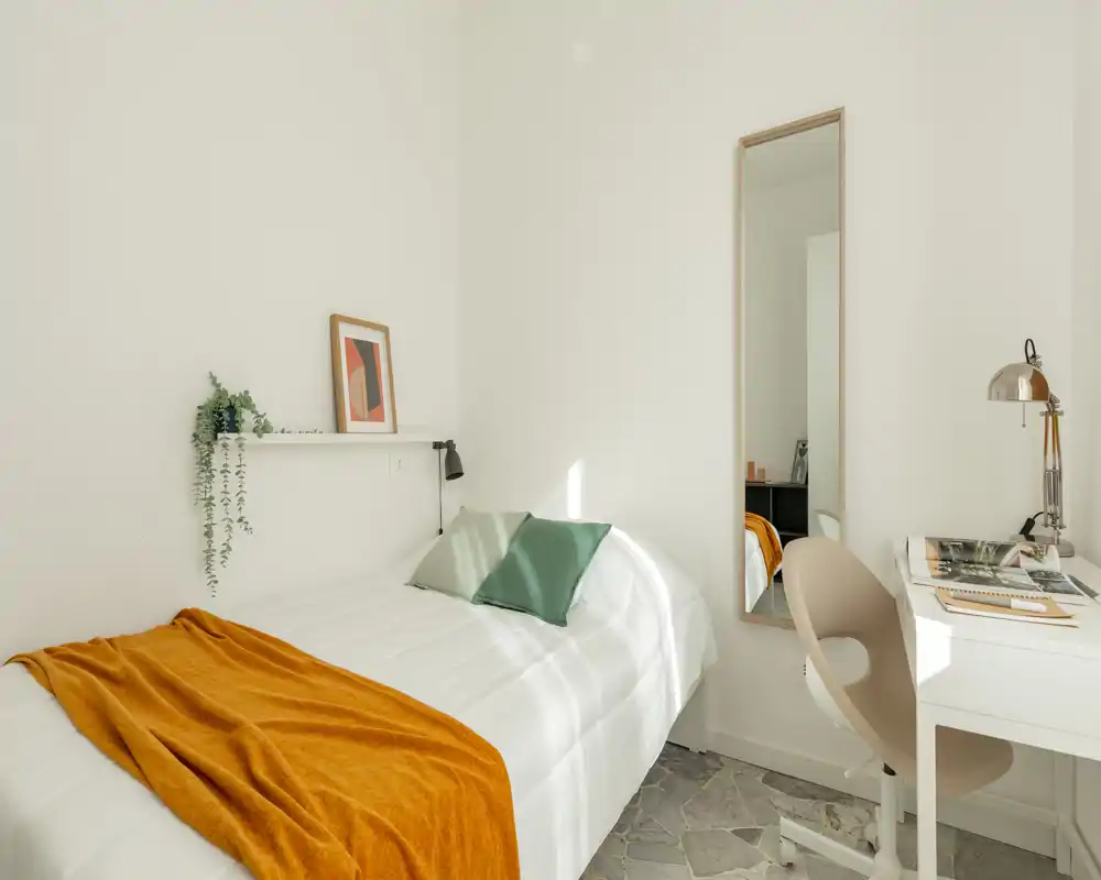 Minimal, sunlit bedroom with a neatly made single bed, warm orange throw and green accent pillows; a slim full-length mirror and a compact white desk create a bright, tidy workspace.