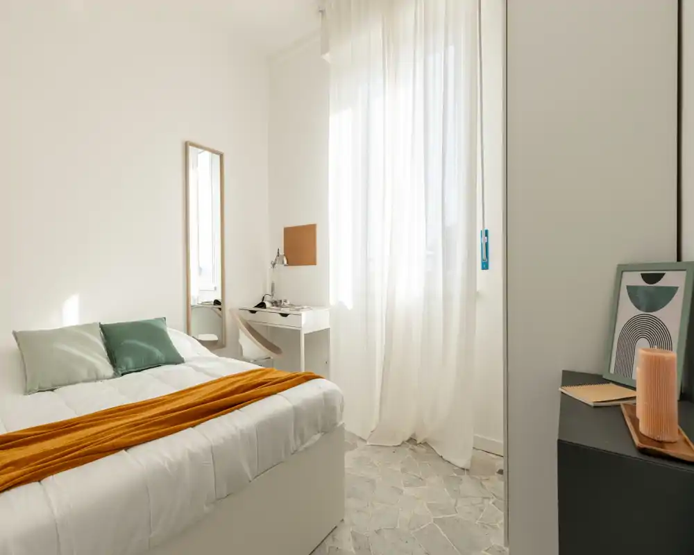 Bright, minimalist bedroom with a neatly made bed, soft cushions and a warm throw, plus a small desk by the window offering natural light.