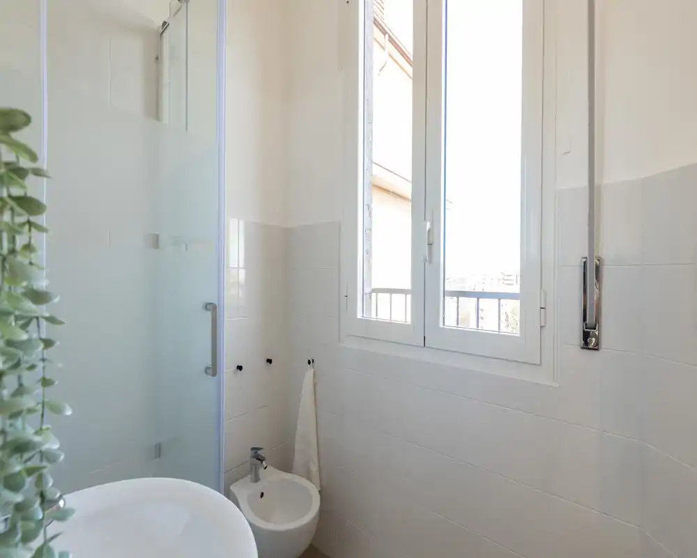 Bagno luminoso e pulito con bidet, box doccia con vetro satinato, lavabo e una finestra che offre luce naturale.