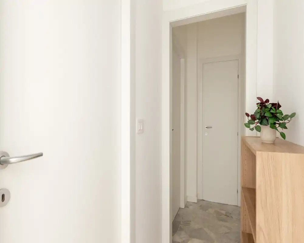 Ingresso luminoso e minimale con mobiletto in legno chiaro e pianta, pareti bianche e pavimento in pietra chiara resistente.
