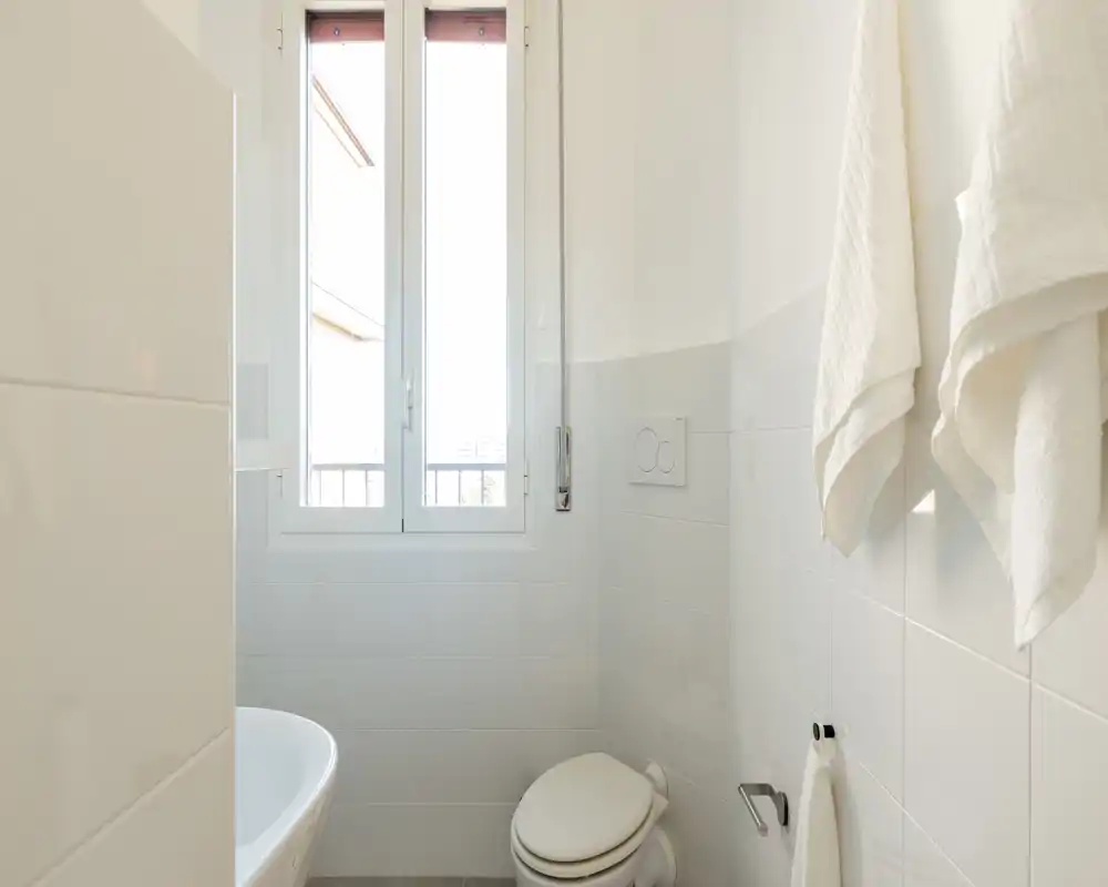 Bagno piccolo e luminoso con WC, portasciugamani e una finestra che illumina l'ambiente; piastrelle bianche donano un aspetto fresco e minimalista.