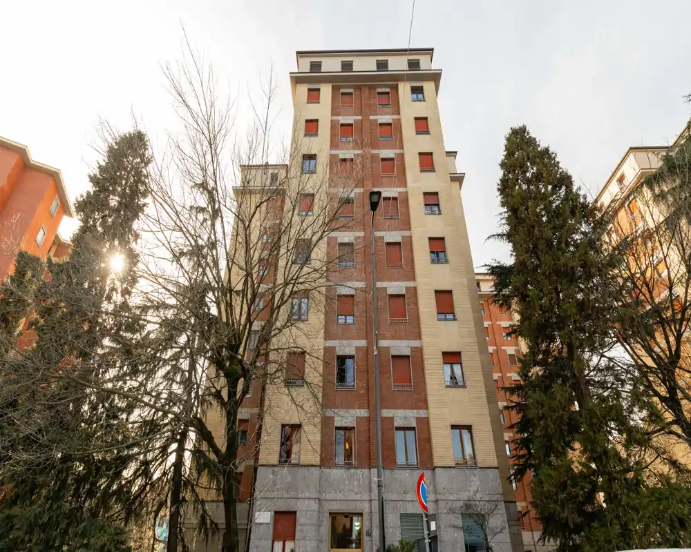 Vista esterna di un condominio multipiano con alberi e un lampione davanti, fotografato con luce del giorno morbida.