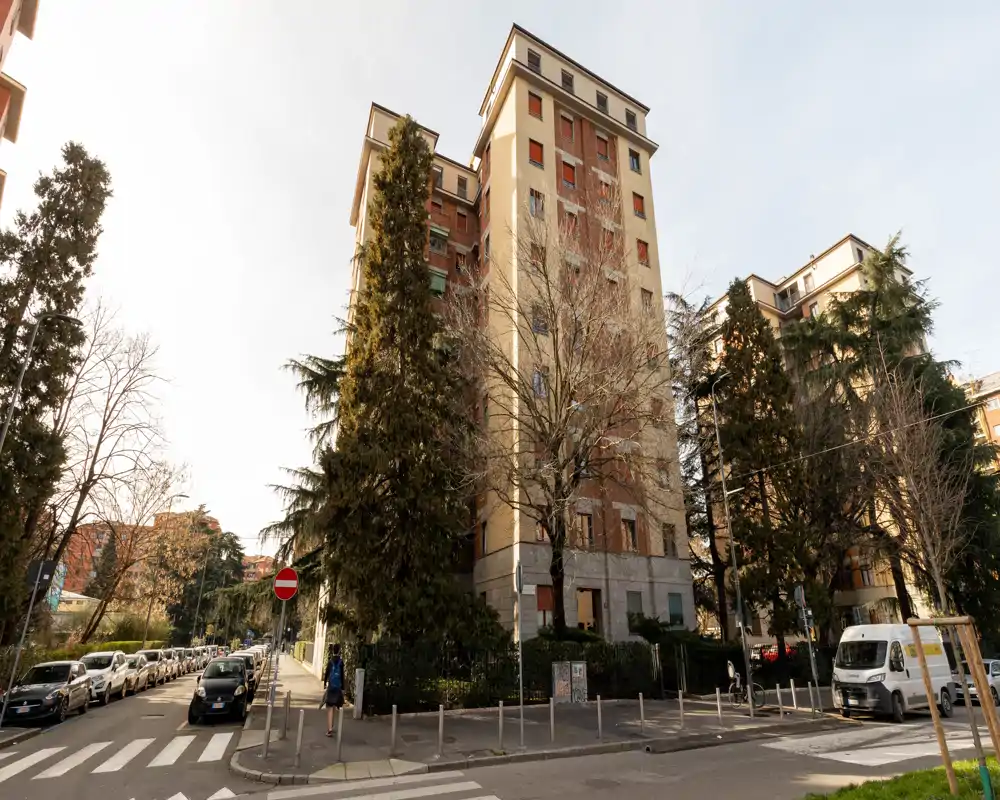 Vista esterna su strada di un edificio residenziale di media altezza con alberi, auto parcheggiate e una striscia pedonale in primo piano.