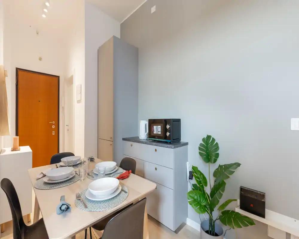 Zona pranzo moderna e luminosa con tavolo apparecchiato per quattro, arredi minimal e pianta decorativa, perfetta per annunci di appartamenti.