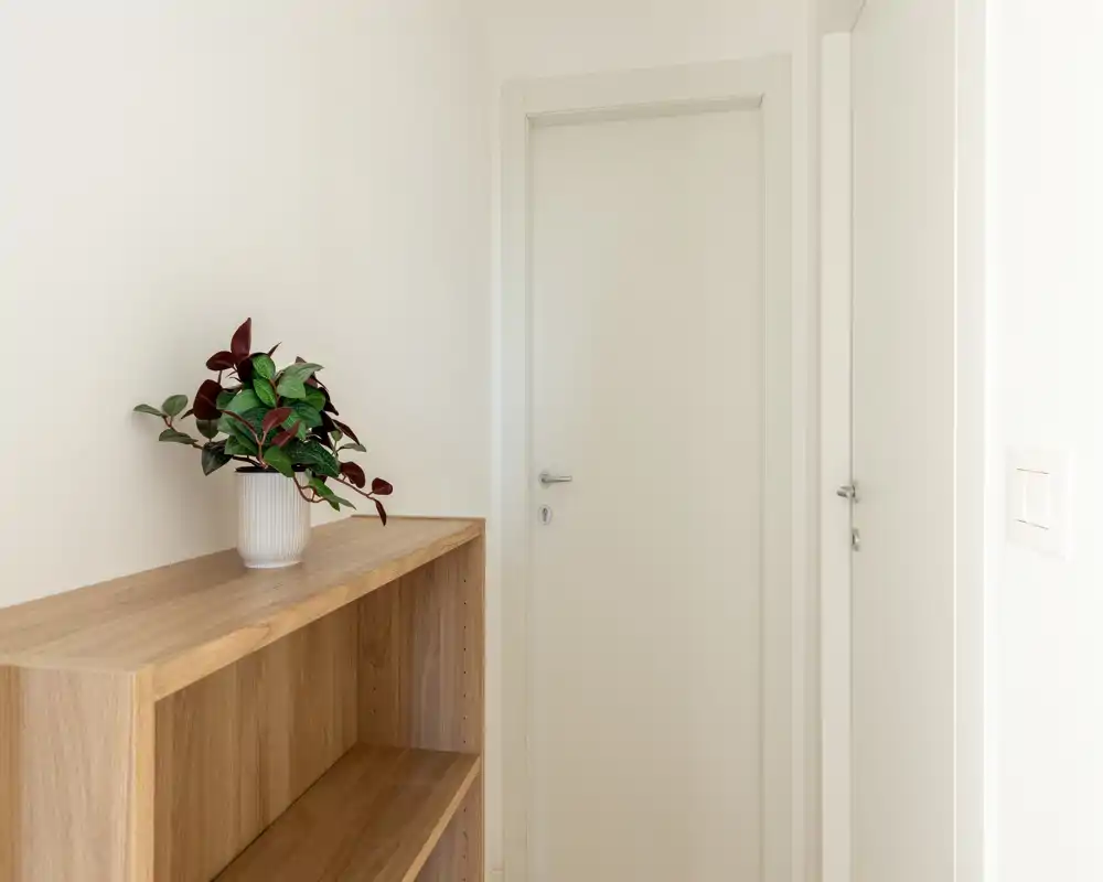 Ingresso/atrio minimale con mobile in legno chiaro e una pianta in vaso, pareti neutre e porta interna chiusa — luminoso, ordinato e accogliente.