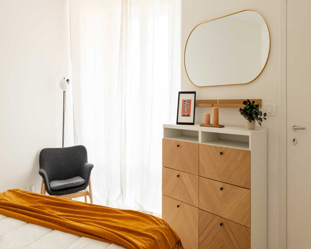 Chambre lumineuse et minimaliste avec un plaid ocre sur le lit, un fauteuil cosy près de voilages et une commode à façades bois surmontée d'un miroir et d'objets décoratifs.