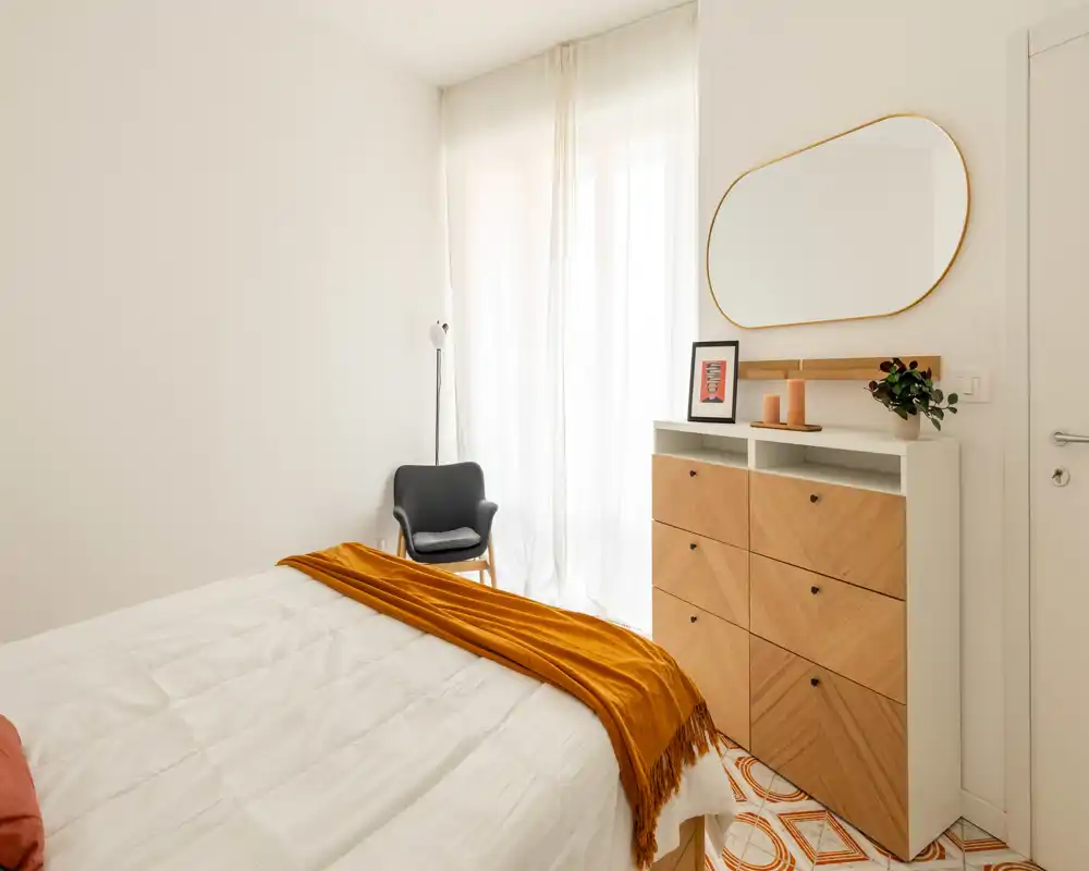 Chambre lumineuse au style doux avec un lit double en linge blanc et un plaid ocre chaleureux. La lumière naturelle traverse des voilages; une commode en bois et un fauteuil apportent une atmosphère minimaliste et accueillante.
