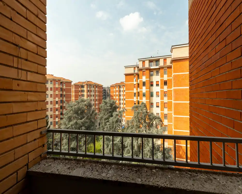 Balcon avec vue sur une cour résidentielle, immeubles en briques orange et arbres matures, encadré par une rambarde métallique et des murs en briques.